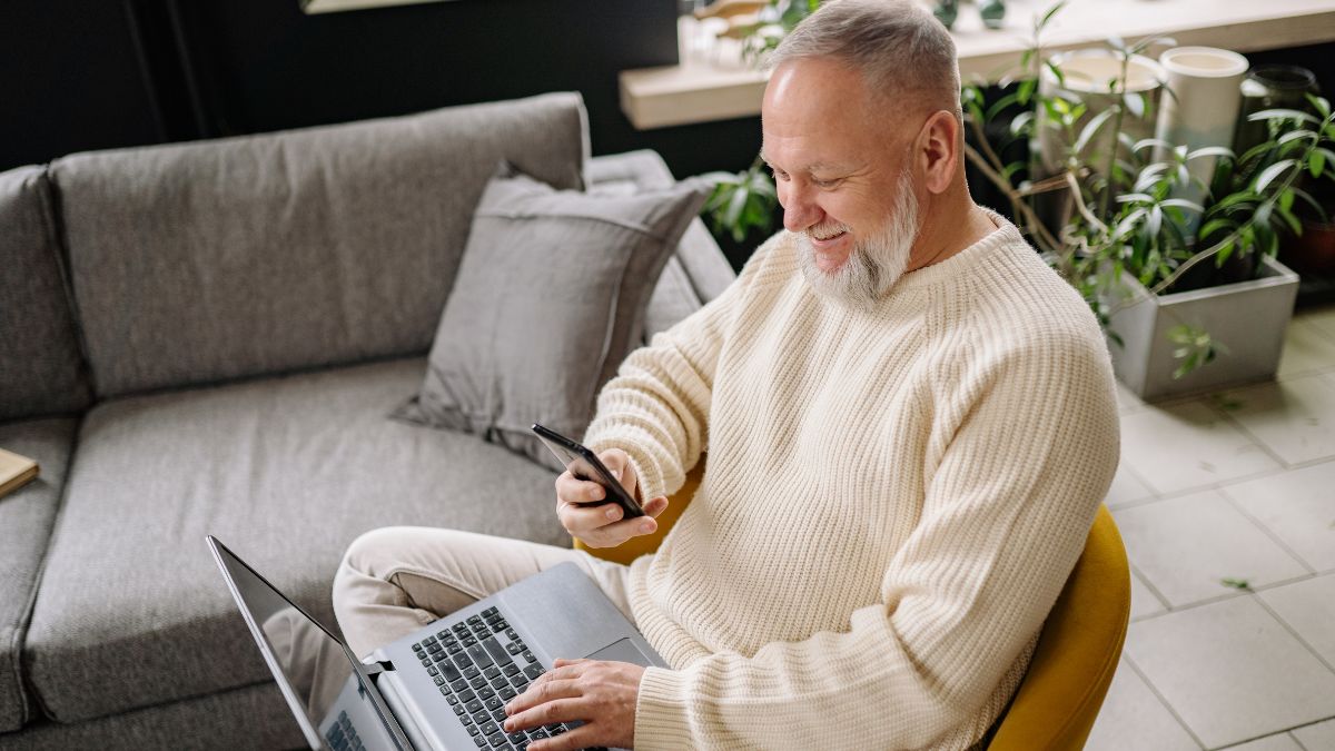 Senior citizen man performs quick email verification on his phone before sending an email on his laptop.