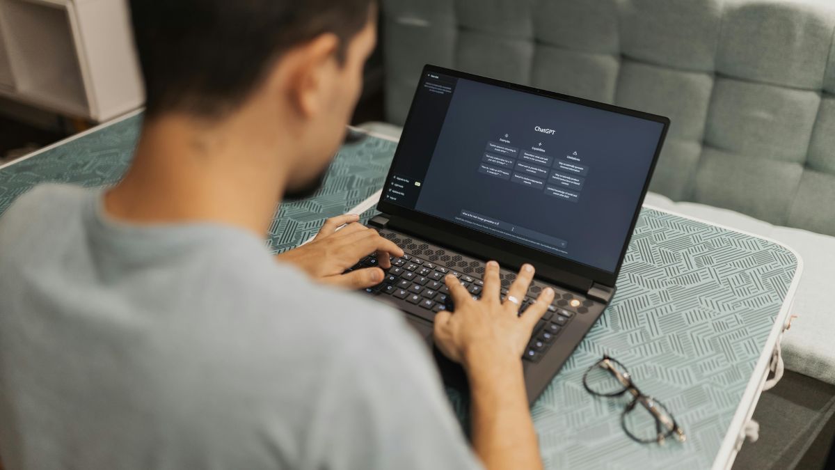 Young man sits in front of a laptop to use ChatGPT for some vibe coding, his spectacles nearby.